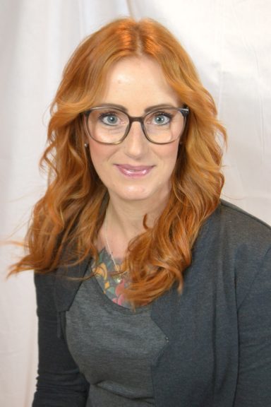Frau mit roten Haaren, Brille und grauem Cardigan, lächelt in die Kamera.