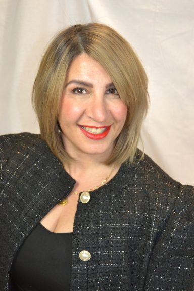 Frau mit blonden, kurzen Haaren, rotem Lippenstift und schwarzem Blazer lächelt.