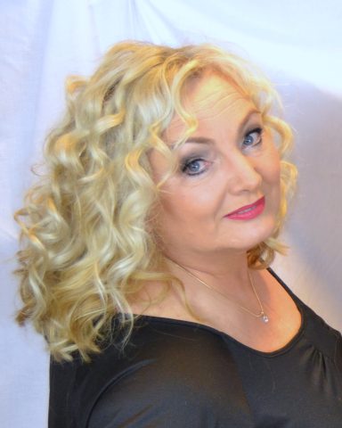 Frau mit lockigem, blondem Haar, lächelt in schwarzem Oberteil vor hellem Hintergrund.