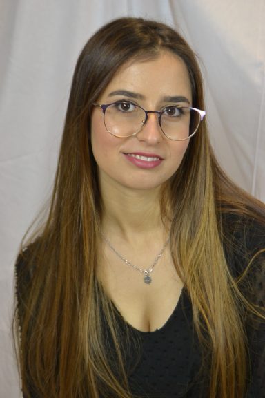 Junge Frau mit langen Haaren und Brille, lächelnd vor einem neutralen Hintergrund.